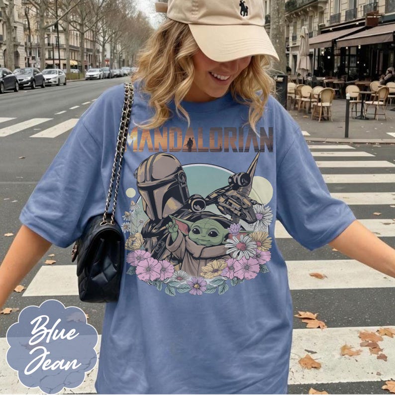 May include: A light blue t-shirt featuring a graphic of The Mandalorian and Grogu surrounded by flowers. The text "MANDALORIAN" is arched across the top. The shirt also has the text "Blue Jean" in a stylized font.
