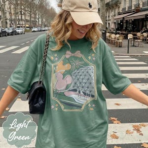 May include: Light green t-shirt with a graphic design featuring a Mickey Mouse balloon, Spaceship Earth, and a monorail. The shirt has short sleeves and a relaxed fit. The words "Light Green" are printed on the bottom left.