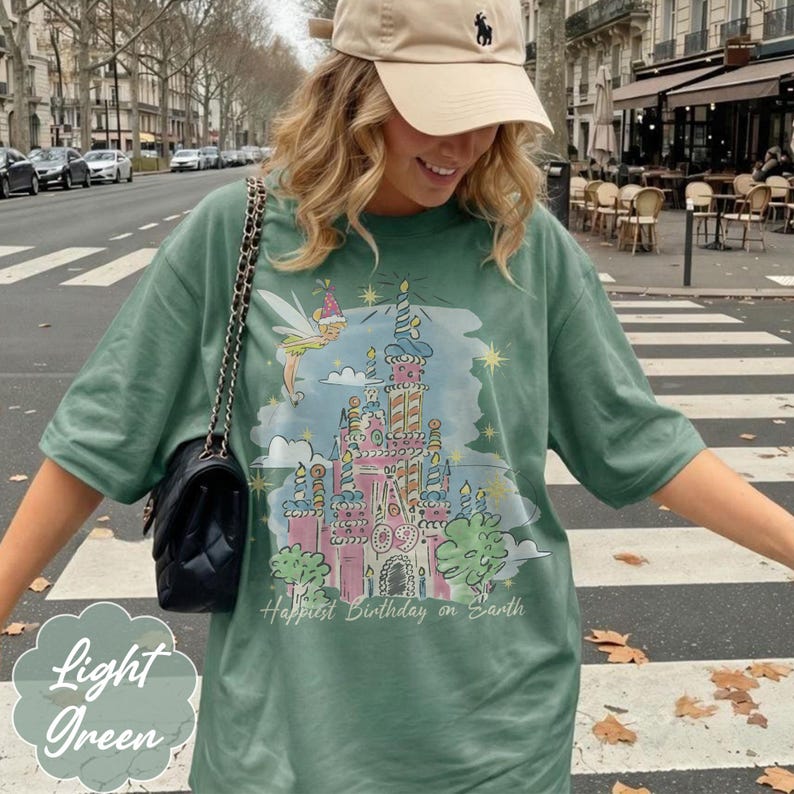 Puede incluir: Camiseta verde claro con un castillo colorido, un hada y el texto "Happiest Birthday on Earth". La camiseta tambi&eacute;n tiene las palabras "Light Green" en una fuente decorativa.