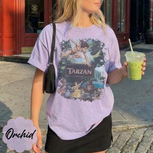 Op de afbeelding: Een lichtpaars T-shirt met een Tarzan-afbeelding met junglekarakters. Het shirt heeft korte mouwen en een ronde hals. Het woord "Tarzan" is in een kader gedrukt. Het wordt gedragen met een zwarte rok en een zwarte schoudertas.