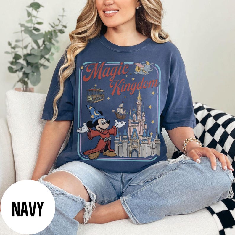 May include: Navy blue t-shirt with a retro graphic of Mickey Mouse as a sorcerer, and the words "Magic Kingdom" above a castle illustration. The shirt has a relaxed fit and is made of a soft material.