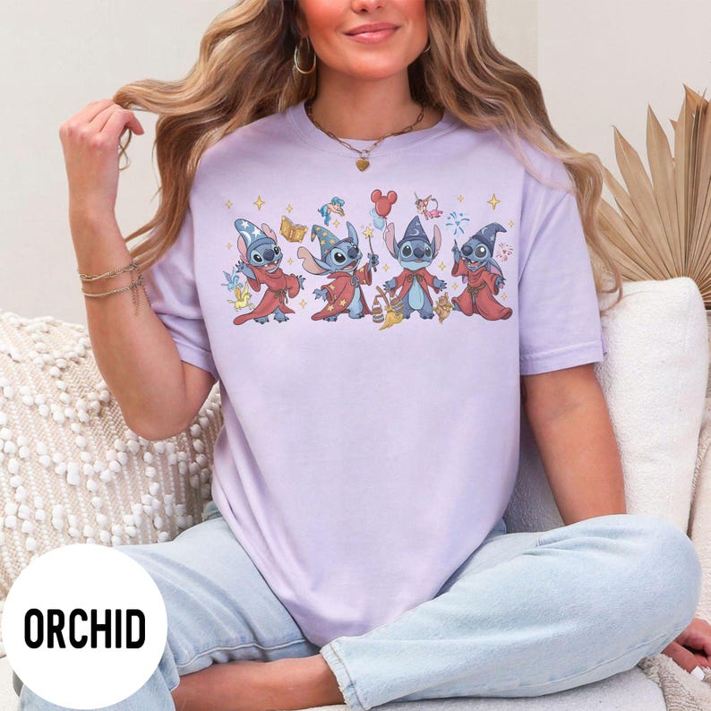 May include: A light purple t-shirt with a graphic of four cartoon characters dressed as wizards. The characters are blue with large ears and are wearing red robes and wizard hats. The word "ORCHID" is printed in a white circle at the bottom.