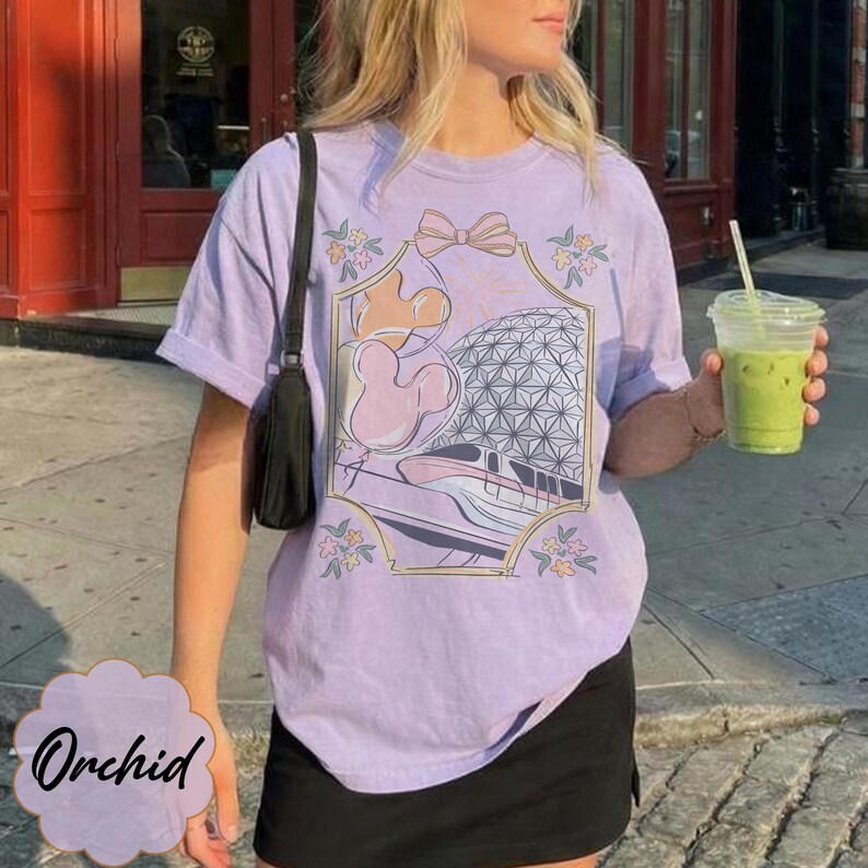 May include: A light purple t-shirt featuring a graphic design with a bow, floral accents, and a stylized image of a theme park. The word "Orchid" is written in a cursive font on a light purple background.