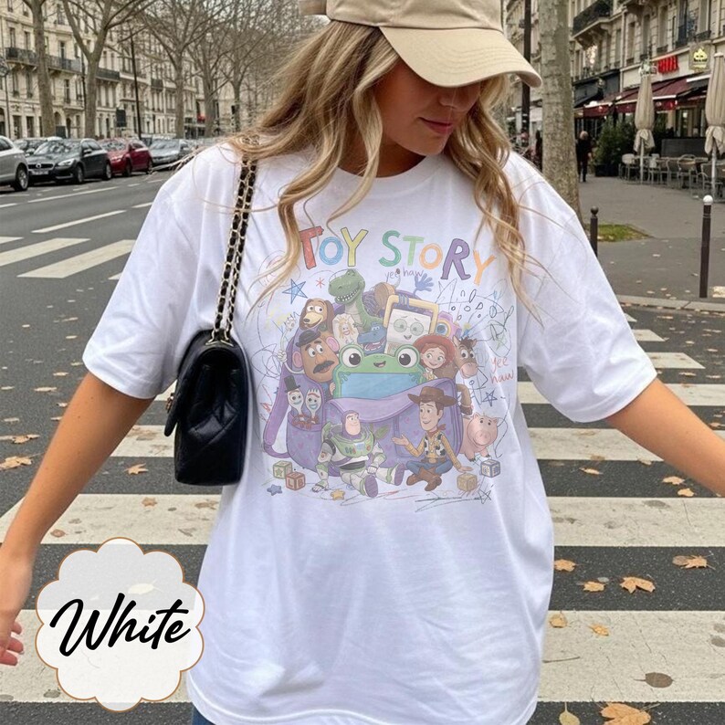 May include: A white short-sleeved t-shirt featuring a colourful graphic print of Toy Story characters including Woody, Buzz Lightyear, and a green frog emerging from a purple backpack, with the text Toy Story printed in multicoloured letters above.