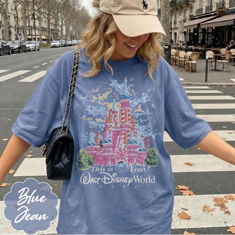 K&ouml;nnte beinhalten: Ein blaues T-Shirt mit einer Grafik eines rosafarbenen Schlosses mit rot-wei&szlig; gestreiften T&uuml;rmen, dem Text 25, This is Now! und Walt Disney World 1997. Das Shirt hat kurze &Auml;rmel, einen Rundhalsausschnitt und wird mit einer schwarzen gesteppten Umh&auml;ngetasche kombiniert.