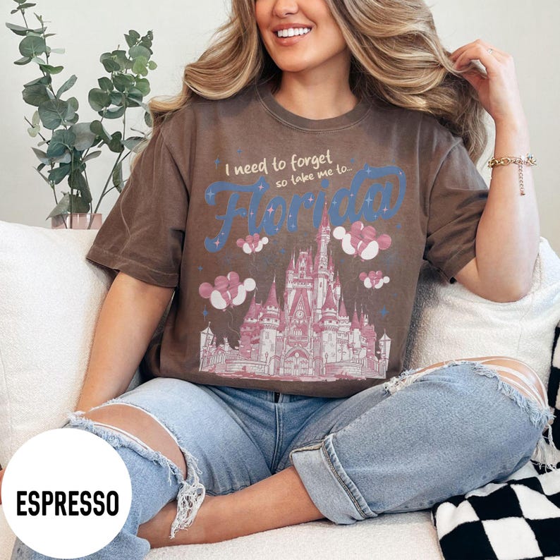 May include: Espresso-colored t-shirt with a graphic of a castle, balloons, and the word "Florida" in blue and white. The text "I need to forget so take me to..." is above the word Florida. The shirt is worn by a person.