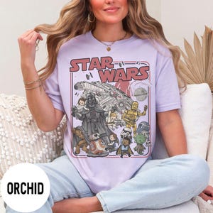 May include: A light purple t-shirt featuring a Star Wars graphic. The graphic includes Darth Vader, C-3PO, R2-D2, Yoda, and other characters, along with the text "STAR WARS" in red. The shirt is labeled "ORCHID" in a white circle.