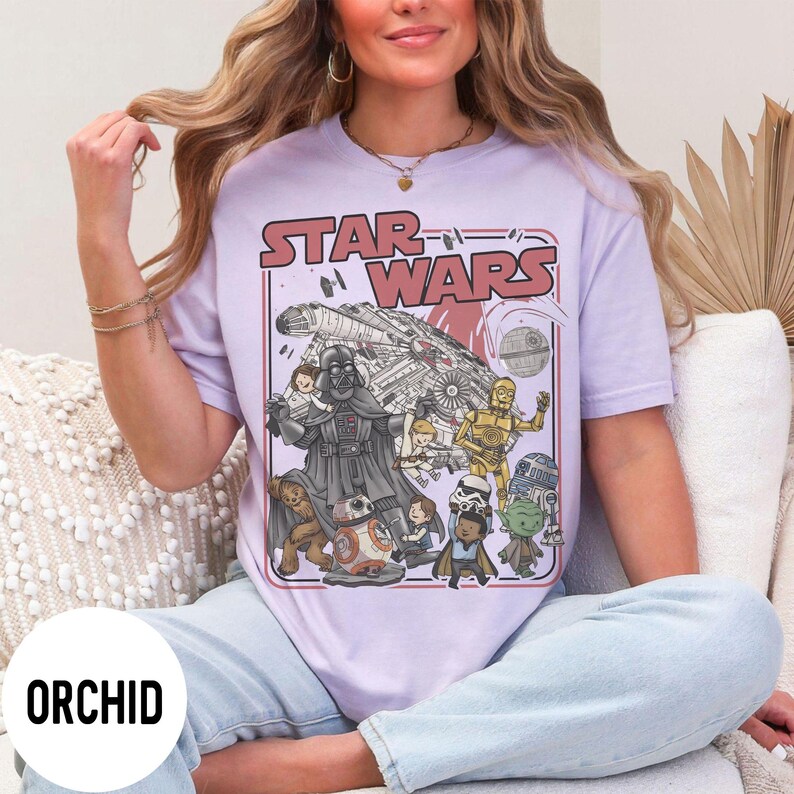 May include: A light purple t-shirt featuring a Star Wars graphic. The graphic includes Darth Vader, C-3PO, R2-D2, Yoda, and other characters, along with the text "STAR WARS" in red. The shirt is labeled "ORCHID" in a white circle.