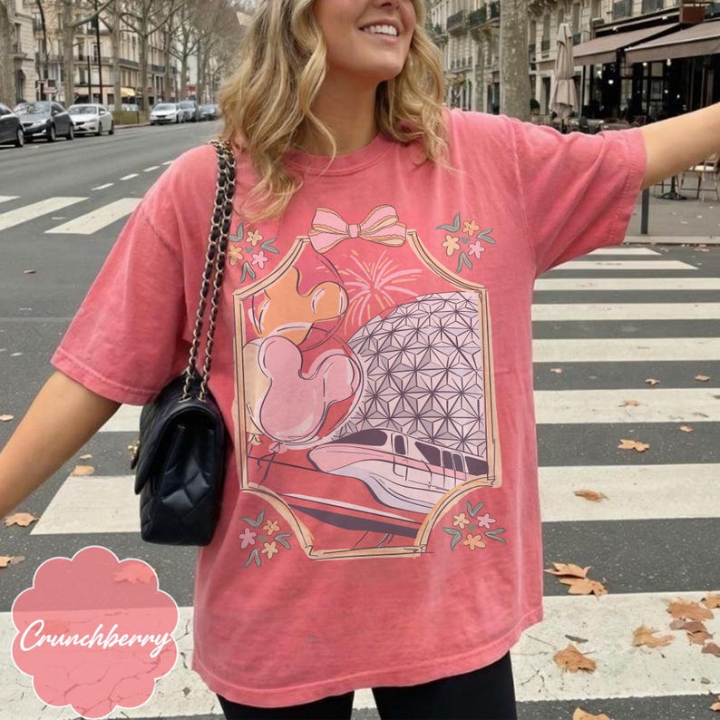 May include: A coral-colored t-shirt featuring a graphic design with a Mickey Mouse head, a geometric sphere, and a monorail. The design is framed with a bow and floral accents. The text "Crunchberry" is at the bottom.
