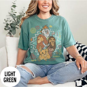 Puede incluir: Camiseta verde claro con un dise&ntilde;o de personajes de El Rey Le&oacute;n. El dise&ntilde;o incluye a Simba, Mufasa, Tim&oacute;n, Pumba y Zaz&uacute;, con patrones coloridos de inspiraci&oacute;n tribal. La camiseta es de corte holgado.