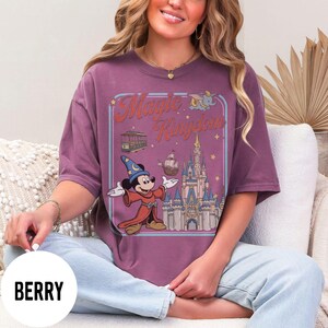 May include: Berry-coloured t-shirt with a graphic of Mickey Mouse as a sorcerer, the words "Magic Kingdom", a castle, and other illustrations. The shirt has a relaxed fit.