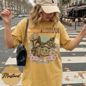 May include: Mustard yellow t-shirt featuring a graphic of The Mandalorian, Grogu, and a spaceship, surrounded by flowers. The word "MANDALORIAN" is printed across the top. The shirt is a relaxed fit.