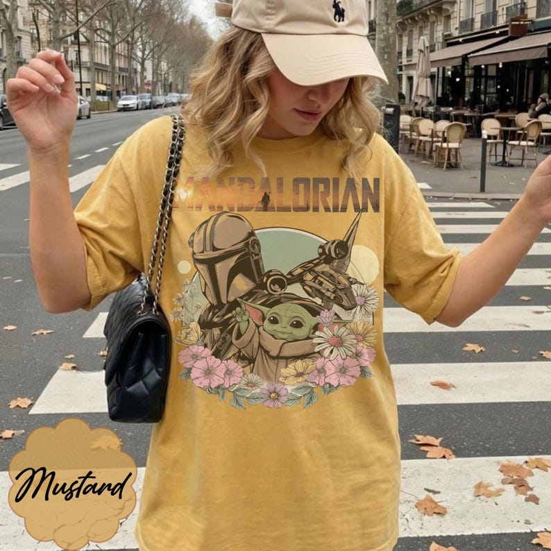 May include: Mustard yellow t-shirt featuring a graphic of The Mandalorian, Grogu, and a spaceship, surrounded by flowers. The word "MANDALORIAN" is printed across the top. The shirt is a relaxed fit.