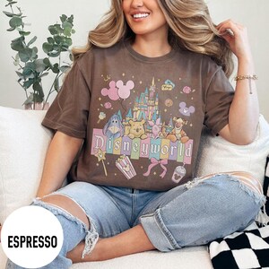 May include: Espresso-colored t-shirt featuring a colorful graphic of Disney characters and the word "Disneyworld." The design includes a castle, balloons, and other themed elements. The shirt is a short-sleeved, casual style.