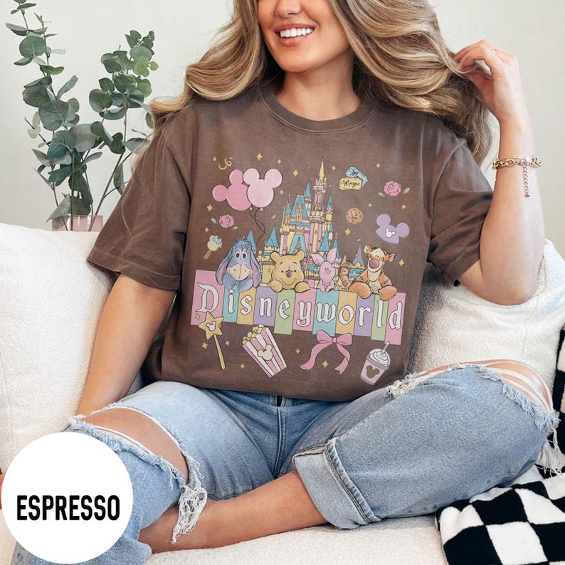 May include: Espresso-colored t-shirt featuring a colorful graphic of Disney characters and the word "Disneyworld." The design includes a castle, balloons, and other themed elements. The shirt is a short-sleeved, casual style.