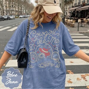 May include: A blue t-shirt featuring a graphic print with the text Cowgirl Like Me and Takes One to Know One. The design includes a red hat, rope motifs, stars, and musical notes. A black quilted shoulder bag with a gold chain strap is worn.