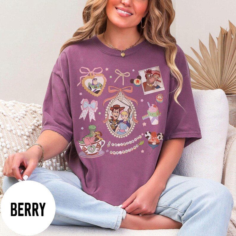 May include: Berry-colored t-shirt featuring cartoon characters and decorative elements. The design includes framed images of characters, bows, and a teacup with a dinosaur. The word "BERRY" is printed on a white circle at the bottom.