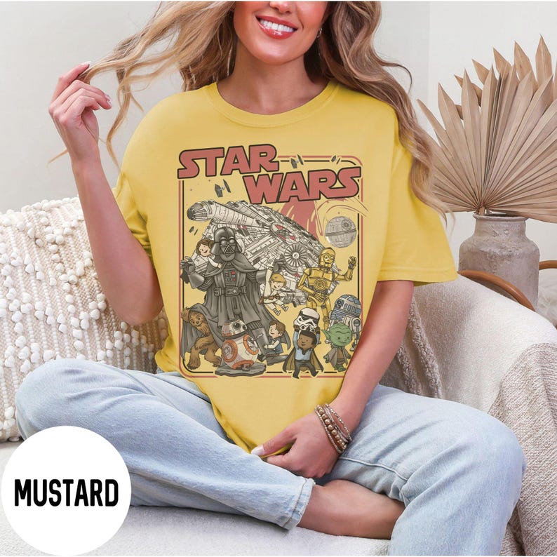May include: Mustard yellow t-shirt featuring a Star Wars graphic. The graphic includes the Star Wars logo, Darth Vader, the Millennium Falcon, and other characters. The shirt is a crew neck and has short sleeves.