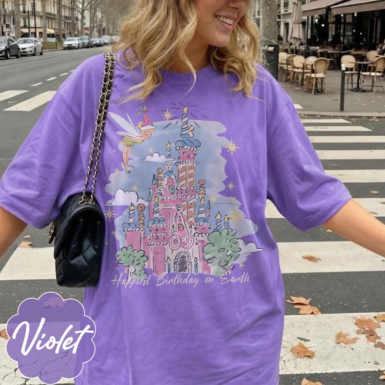 Puede incluir: Camiseta violeta con un dise&ntilde;o de castillo de cuento de hadas, un hada y el texto "Happiest Birthday on Earth". El gr&aacute;fico utiliza colores pastel y estrellas. La camiseta tiene un corte holgado.