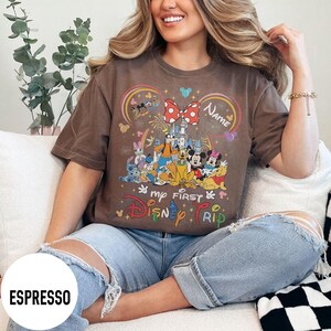 May include: Espresso-colored t-shirt featuring a colorful Disney graphic with Mickey and Minnie Mouse, and other characters. The text "my FIRST Disney TRIP" is displayed below the graphic. The shirt has a relaxed fit.