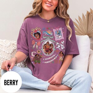 Pu&ograve; includere: T-shirt color bacca con un design grafico colorato. Il design include ritratti di personaggi incorniciati, fiocchi e un ritratto ovale centrale di una famiglia. La parola "BERRY" &egrave; stampata in un cerchio bianco in basso.