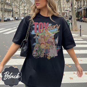 May include: A black short-sleeved t-shirt featuring a graphic print with the text "TOY story" and an illustration of various animated characters in colorful outfits. The shirt has a crew neckline and a loose, relaxed fit with short sleeves.