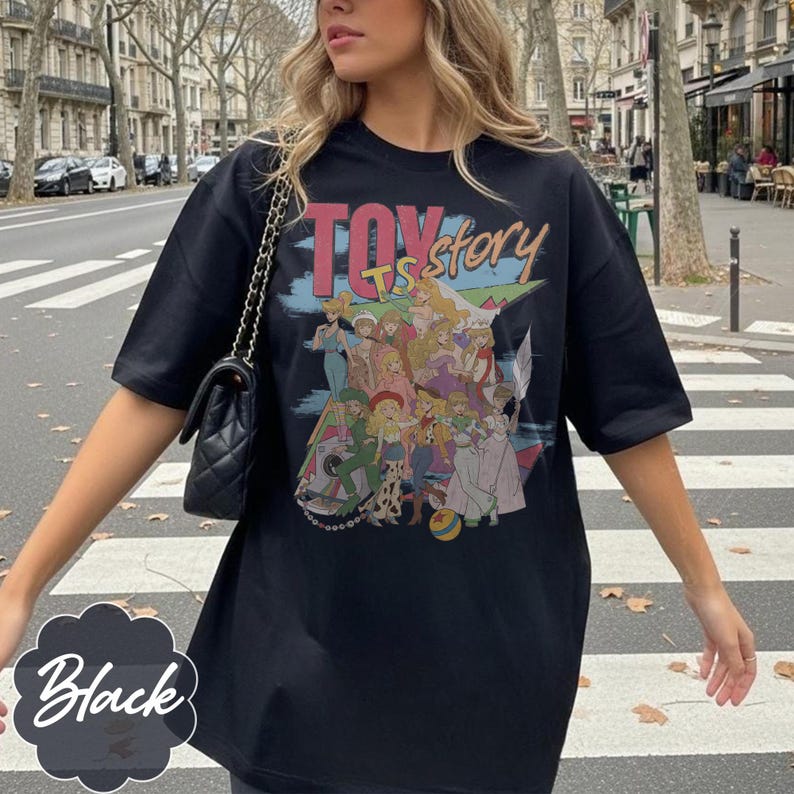 May include: A black short-sleeved t-shirt featuring a graphic print with the text "TOY story" and an illustration of various animated characters in colorful outfits. The shirt has a crew neckline and a loose, relaxed fit with short sleeves.