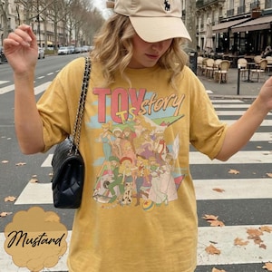 May include: A mustard yellow short-sleeve t-shirt featuring a graphic print of various Toy Story characters. The text "TOY story" is printed in large red and orange letters above the illustrated group of characters, which includes Bo Peep.
