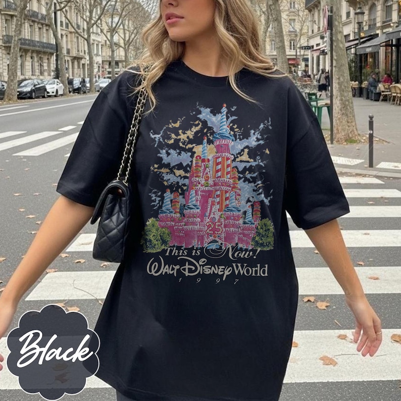 K&ouml;nnte beinhalten: Ein schwarzes Kurzarm-T-Shirt mit einer Grafik eines rosafarbenen Schlosses mit blauen Akzenten, dem Text "This is Now!", "Walt Disney World" und "1997". Das Design enth&auml;lt ein "25"-Emblem am Schloss und eine schwarze gesteppte Umh&auml;ngetasche.