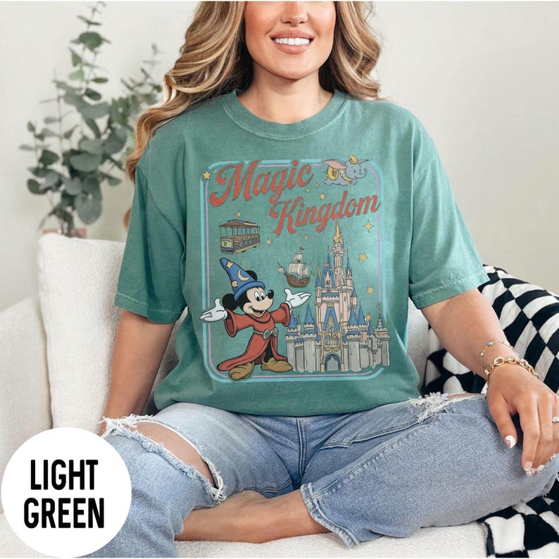 May include: Light green t-shirt featuring a graphic of Mickey Mouse in a sorcerer's outfit in front of a castle. The words "Magic Kingdom" are in a frame above the image. The shirt is a light green colour.