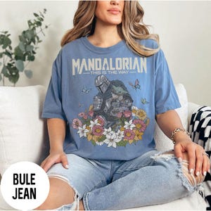Puede incluir: Camiseta azul claro con un dise&ntilde;o de The Mandalorian y Grogu rodeados de flores y mariposas. El texto "MANDALORIAN - THIS IS THE WAY" est&aacute; impreso en la parte superior. La camiseta es de material suave.