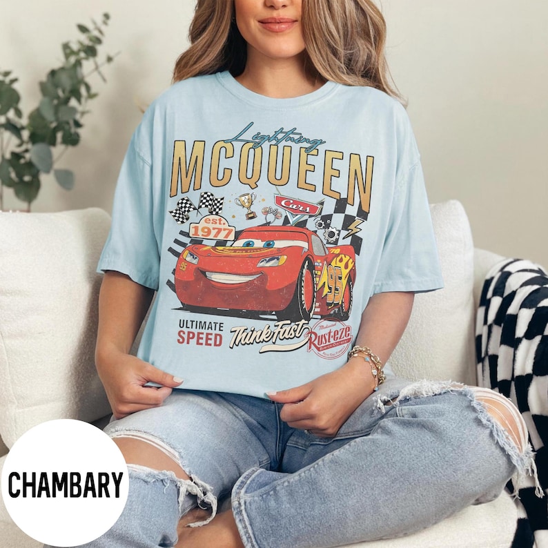 Op de afbeelding: Lichtblauw T-shirt met een afbeelding van Lightning McQueen uit de film Cars. Het ontwerp bevat de tekst "Lightning McQueen" en "Cars est. 1977" met racevlaggen en een trofee. Het shirt heeft ook de zinnen "Ultimate Speed" en "Think Fast".