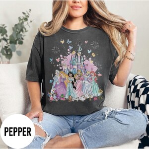 May include: Dark gray t-shirt featuring a colorful graphic of Disney princesses in front of a castle. The graphic includes fireworks, butterflies, and Mickey Mouse balloons. The word "PEPPER" is printed in white on the bottom left.
