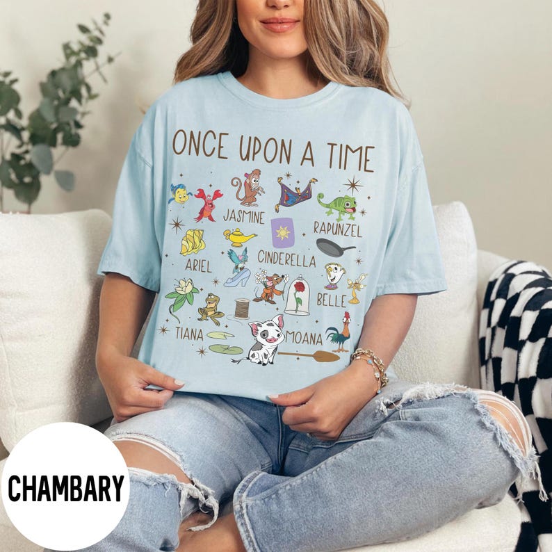 May include: Pale blue t-shirt featuring the phrase "ONCE UPON A TIME" and cartoon illustrations of Disney characters. The design includes characters such as Ariel, Jasmine, Cinderella, and Moana. The t-shirt is a light blue colour.