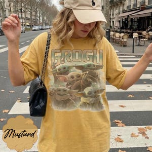 May include: A mustard yellow short-sleeve t-shirt featuring a graphic print of four small green creatures with large ears and eyes. The text GROGU is printed in a bold, stylized font above the creatures. The shirt has a crew neckline and loose fit.