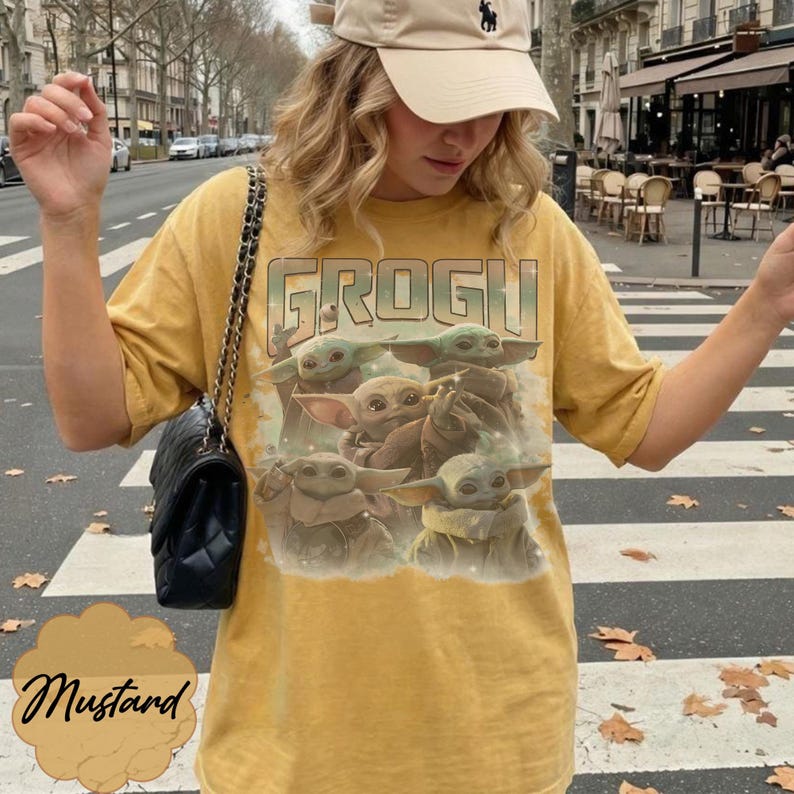 May include: A mustard yellow short-sleeve t-shirt featuring a graphic print of four small green creatures with large ears and eyes. The text GROGU is printed in a bold, stylized font above the creatures. The shirt has a crew neckline and loose fit.