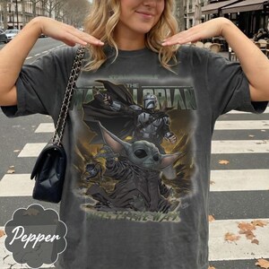 Puede incluir: Una camiseta gris carb&oacute;n de manga corta con un estampado gr&aacute;fico de The Mandalorian y Grogu. El texto dice THE MANDALORIAN en la parte superior y THIS IS THE WAY en la parte inferior. El dise&ntilde;o incluye una capa negra, un bl&aacute;ster y una rana.