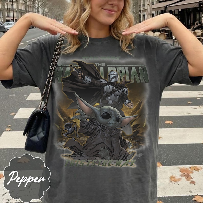 Puede incluir: Una camiseta gris carb&oacute;n de manga corta con un estampado gr&aacute;fico de The Mandalorian y Grogu. El texto dice THE MANDALORIAN en la parte superior y THIS IS THE WAY en la parte inferior. El dise&ntilde;o incluye una capa negra, un bl&aacute;ster y una rana.