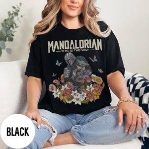 Puede incluir: Camiseta negra con el texto "MANDALORIAN - THIS IS THE WAY" sobre una ilustraci&oacute;n de un personaje y Grogu rodeados de flores y mariposas.