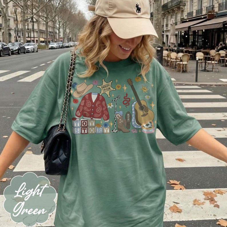 May include: A sage green t-shirt featuring a graphic print of a cowboy hat, a red paisley cardigan, a sheriff badge labeled Sheriff Taylor, a microphone, a brown cowboy boot, a guitar, and blocks spelling Toy Story with cow print accents.