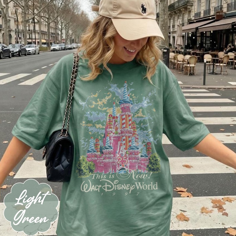 K&ouml;nnte beinhalten: Ein salbeigr&uuml;nes Kurzarm-T-Shirt mit einer Grafik eines rosafarbenen Schlosses mit blauen Akzenten, der Zahl 25 und dem Text This is Now! Walt Disney World 1997. Dazu geh&ouml;rt eine schwarze, gesteppte Umh&auml;ngetasche mit einer goldfarbenen Kette.