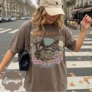 May include: Espresso-colored t-shirt featuring a graphic of The Mandalorian and Grogu surrounded by flowers. The shirt has short sleeves and a crew neck. The word "Mandalorian" is printed across the top of the graphic.