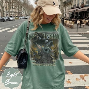 Puede incluir: Una camiseta verde salvia con un estampado gr&aacute;fico de The Mandalorian y Grogu. El texto dice The Mandalorian en la parte superior y This Is The Way en la parte inferior. La camiseta tiene mangas cortas y cuello redondo, con un bolso negro acolchado.