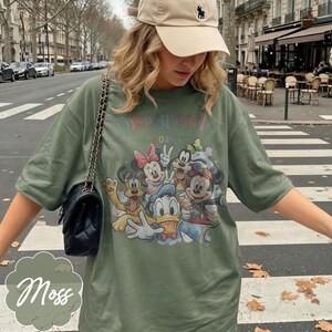 Pu&ograve; includere: T-shirt verde muschio con grafica di personaggi Disney, tra cui Topolino, Minnie e Paperino. Il testo "Disneyworld 2026" &egrave; stampato sopra i personaggi. La maglietta ha una vestibilit&agrave; ampia.