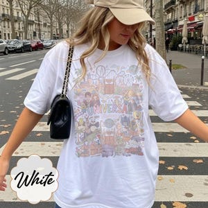 May include: White t-shirt with a colourful graphic featuring the text "Universal Studios" and various cartoon characters and theme park elements. The shirt is a casual style, ideal for a day out.