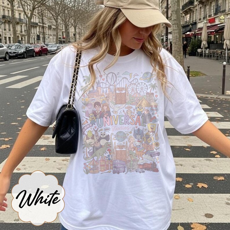 May include: White t-shirt with a colourful graphic featuring the text "Universal Studios" and various cartoon characters and theme park elements. The shirt is a casual style, ideal for a day out.
