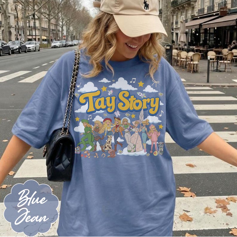 May include: A blue jean colored t-shirt featuring a graphic print with the text Tay Story in yellow letters. The design includes illustrations of various figures in different outfits against a background of white clouds, yellow stars, and musical notes.