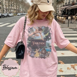 Op de afbeelding: Lichtroze T-shirt met een Tarzan-afbeelding. De afbeelding bevat het woord "Tarzan" op een houten bord, met afbeeldingen van Tarzan, Jane en andere personages. Het shirt heeft een casual stijl.