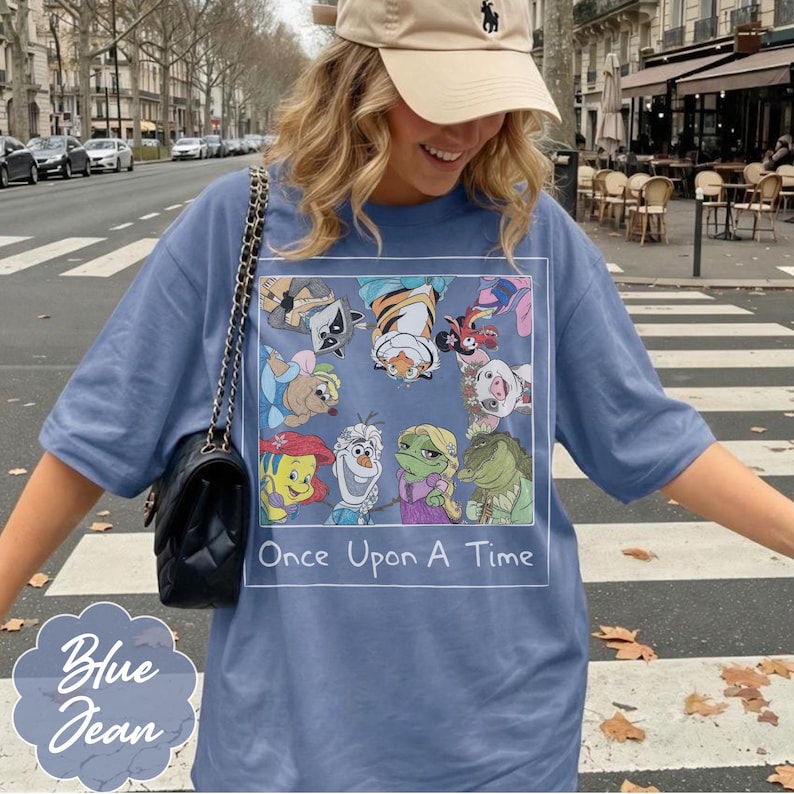 May include: Blue jean t-shirt featuring a colorful graphic of cartoon characters with the text "Once Upon A Time" in a white rectangle. The shirt has short sleeves and a relaxed fit. A light blue logo that says "Blue Jean" is in the bottom left corner.