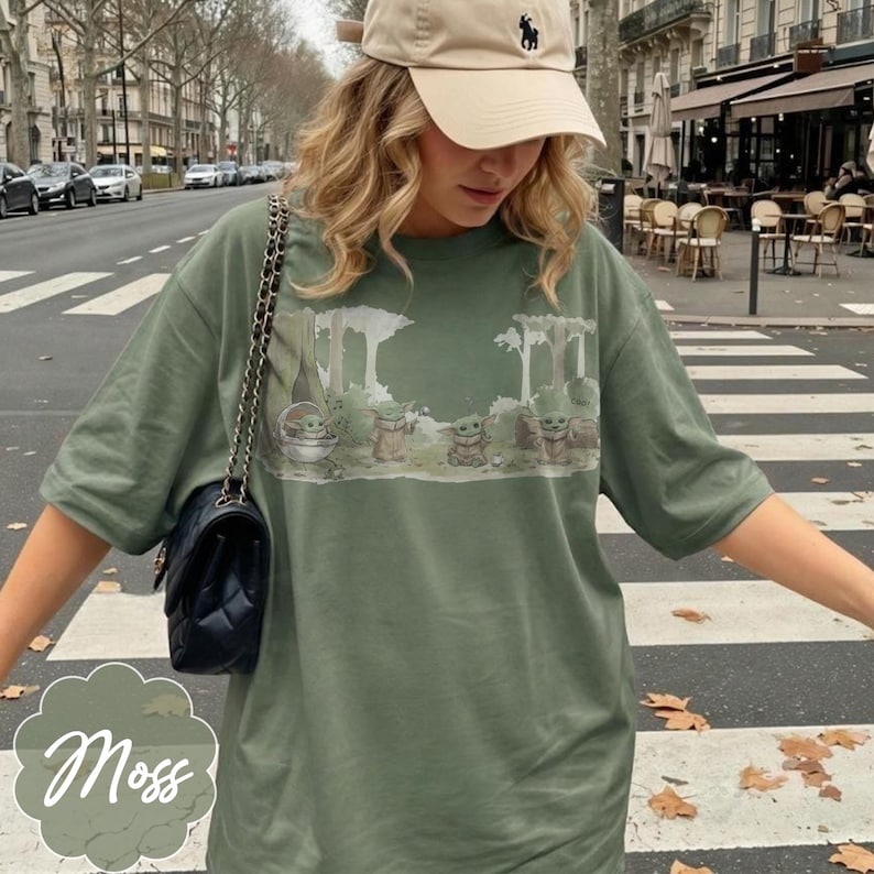 Puede incluir: Camiseta verde musgo con un estampado de escena de bosque con varios personajes de Grogu. La camiseta tiene mangas cortas y un corte holgado. La palabra "Moss" est&aacute; escrita en cursiva en la esquina inferior izquierda.